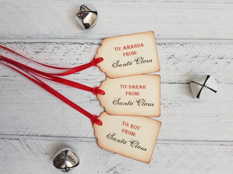 Puede incluir: Tres etiquetas de regalo de estilo vintage con cinta roja. Las etiquetas son marrones con un aspecto desgastado y tienen el texto "TO: AMANDA FROM: Santa Claus", "TO: SARAH FROM: Santa Claus" y "TO: ROY FROM: Santa Claus" impreso en rojo.