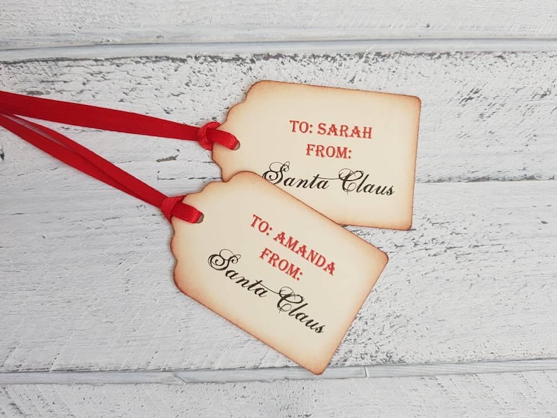 Puede incluir: Dos etiquetas de regalo de estilo vintage con cinta roja. Las etiquetas son de color crema con bordes marrones y tienen el texto "TO: SARAH FROM: Santa Claus" y "TO: AMANDA FROM: Santa Claus" escrito en cursiva negra.