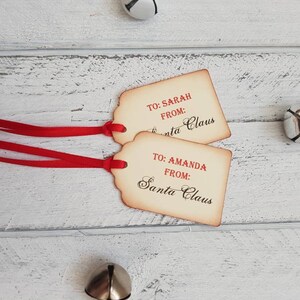 Puede incluir: Dos etiquetas de regalo de estilo vintage con cinta roja. Las etiquetas son marrones con texto blanco que dice "TO: SARAH FROM: Santa Claus" y "TO: AMANDA FROM: Santa Claus".