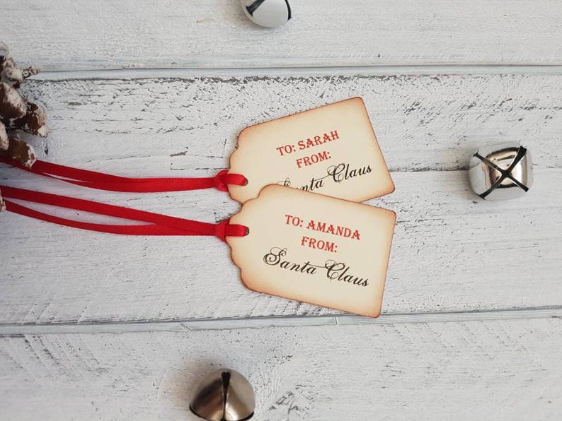 Puede incluir: Dos etiquetas de regalo de estilo vintage con cinta roja. Las etiquetas son marrones con texto blanco que dice "TO: SARAH FROM: Santa Claus" y "TO: AMANDA FROM: Santa Claus".