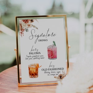 May include: A gold-framed sign with a floral design and the text "Signature Drinks". The sign lists two drinks: "hers PALOMA Tequila, grapefruit juice, lime juice, sparkling water" and "his OLD FASHIONED Whiskey, bitters, sugar, ice, orange peel".