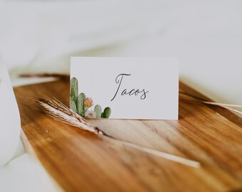 May include: A white card with the word "Tacos" written in black calligraphy. The card has a green cactus and orange flower design in the bottom left corner. The card is sitting on a wooden surface.