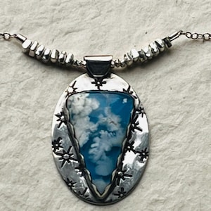 May include: A silver pendant necklace featuring an oval-shaped pendant with a blue and white stone center, resembling a cloudy sky. The pendant is framed by a textured silver border with star-like accents. The necklace has a delicate silver chain.