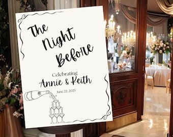 May include: A sign with the text "The Night Before" celebrating Annie & Keith, June 22, 2025. The sign features a drawing of champagne pouring into stacked glasses. The sign is on a brown pedestal.