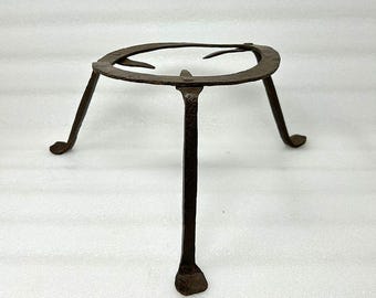 May include: A rustic, dark brown metal trivet with three legs. The trivet has a circular top with three curved supports. The legs are straight and end in small, flat feet. This trivet is likely used for holding hot pots or pans.