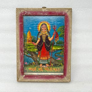 May include: A framed religious print featuring a woman with a halo, holding a trident, standing on a crocodile. The print has a blue background with a temple and river scene. The frame is a distressed wood with red accents.