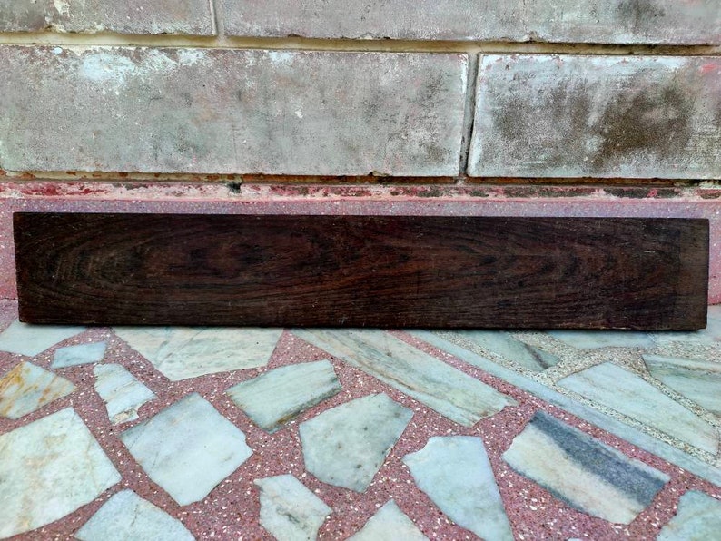 May include: A dark brown wooden plank with a smooth surface. The plank is lying on a tiled floor.