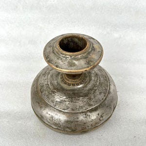 May include: A silver and brass candlestick with a round base and a circular top. The candlestick has a worn, aged appearance.