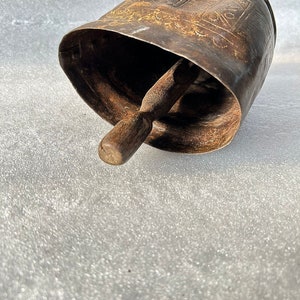 May include: A large, antique, metal cowbell with a wooden clapper. The bell is decorated with an intricate, embossed design.