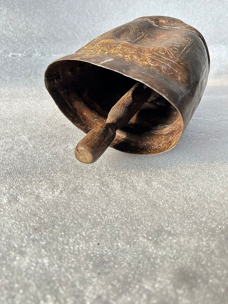May include: A large, antique, metal cowbell with a wooden clapper. The bell is decorated with an intricate, embossed design.