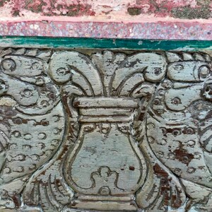 May include: A close-up of a carved wooden panel with a decorative design. The panel is painted in a light gray color and features a central urn with floral motifs on either side. The urn is surrounded by a border of green paint.