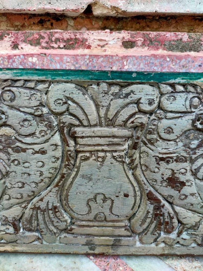 May include: A close-up of a carved wooden panel with a decorative design. The panel is painted in a light gray color and features a central urn with floral motifs on either side. The urn is surrounded by a border of green paint.