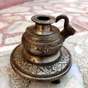 May include: A metallic candle holder with a dark, weathered finish and a circular base. The object features intricate floral engravings on the wide bottom rim and a smaller, patterned mid-section. A small, curved handle is attached to the side.
