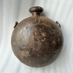 May include: A vintage, round, metal water bottle with a hinged lid and a carrying handle. The bottle is brown and shows signs of wear and age.