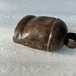 May include: A brown metal cowbell with intricate etched designs. The bell has a worn, aged appearance and a small handle.