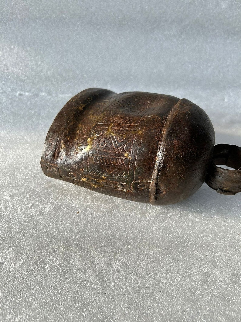 May include: A brown metal cowbell with intricate etched designs. The bell has a worn, aged appearance and a small handle.