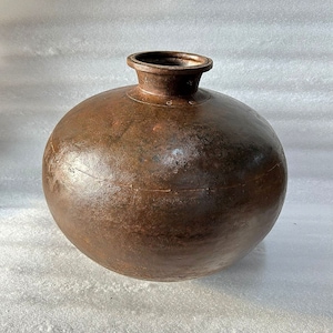 May include: A large, round, brown metal pot with a wide opening at the top. The pot has a slightly textured surface and appears to be made of iron or copper.