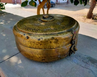Puede incluir: Un recipiente redondo de latón antiguo con tapa. El recipiente tiene un patrón detallado en la tapa y un cierre en el lateral. El latón tiene una pátina cálida y envejecida. El recipiente está sobre una superficie de hormigón.