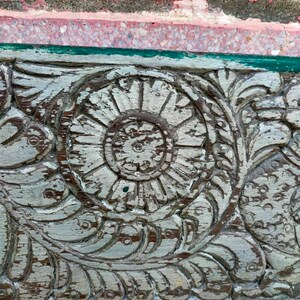 May include: Close-up of a weathered wooden panel with intricate carvings. The carvings feature a central flower design with a circular pattern and surrounding floral motifs. The wood is painted white with areas of the original brown wood showing through.