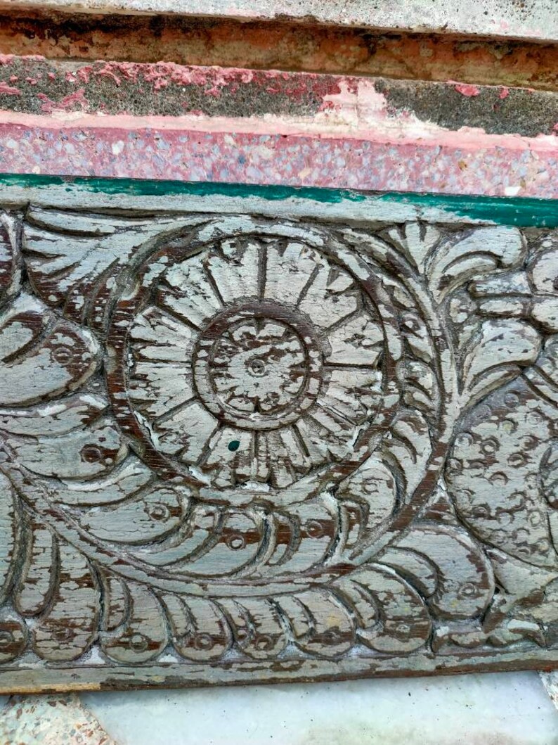 May include: Close-up of a weathered wooden panel with intricate carvings. The carvings feature a central flower design with a circular pattern and surrounding floral motifs. The wood is painted white with areas of the original brown wood showing through.