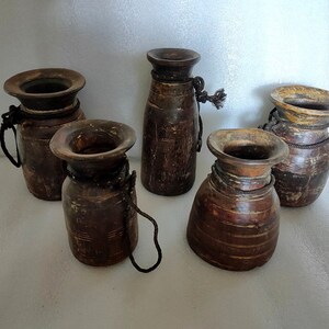 May include: Five rustic, wooden vases with flared rims and dark brown tones. Each vase has a unique shape and is adorned with a dark rope detail around the neck. The vases vary in height and width, showcasing a natural, aged aesthetic.