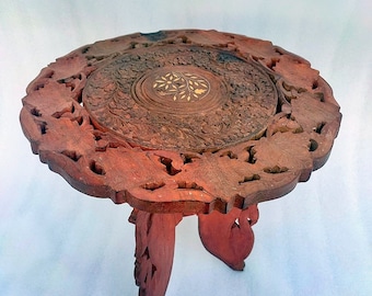 May include: A small, intricately carved wooden table with a dark reddish-brown finish. The round tabletop features detailed floral and leaf patterns, with a central inlaid design. The table stands on three carved legs.