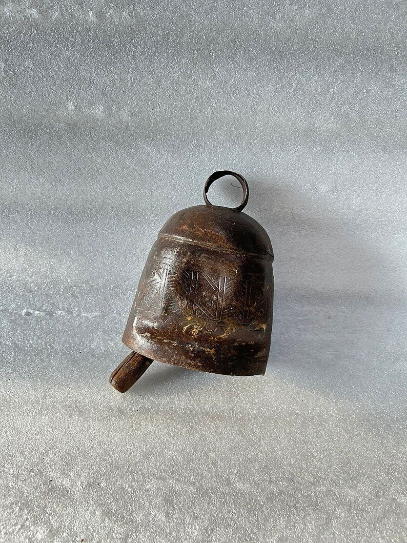 May include: A brown metal bell with a wooden handle and intricate carvings. The bell has a circular loop at the top for hanging.