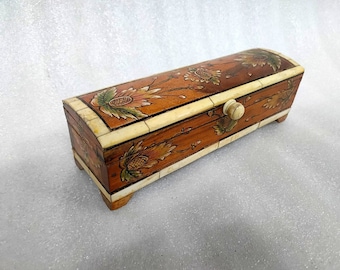 May include: A rectangular wooden box with a hinged lid, featuring hand-painted floral motifs in shades of orange, green, and brown. The box is accented with cream-coloured inlay panels along the edges and includes a small, round wooden knob.