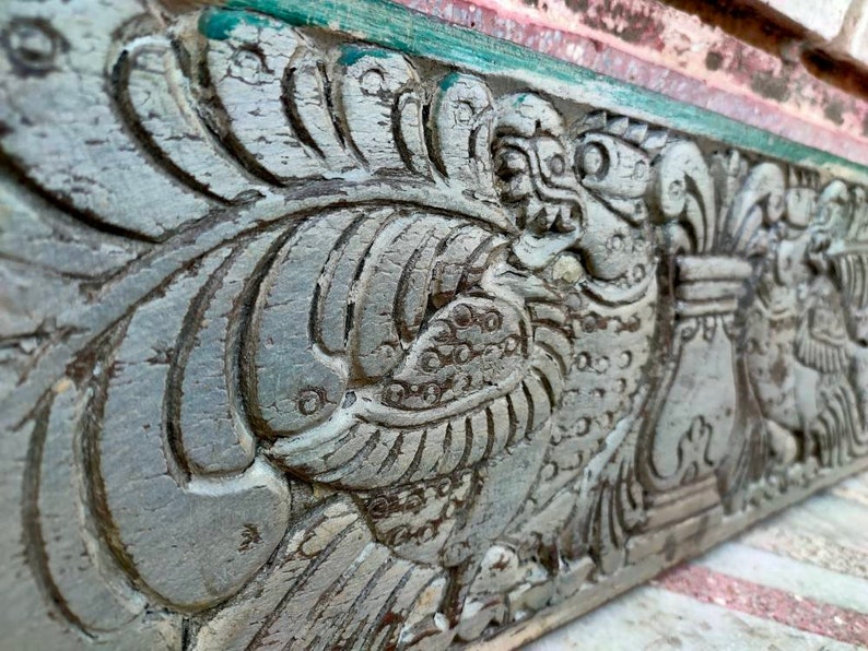 May include: Close-up of a weathered, gray wooden carving with intricate details of a mythical creature with a beak and feathers. The carving is painted with shades of green and white.