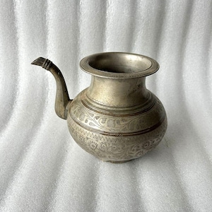 May include: Antique silver-colored metal pitcher with a spout and a wide, round opening. The body of the pitcher is adorned with intricate etched floral and geometric patterns. The handle is curved and detailed.
