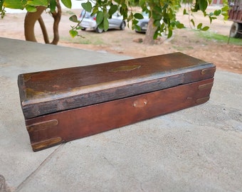May include: A rectangular wooden box with a dark brown finish, featuring brass corner reinforcements and a central brass inlay on the lid. The box has a hinged lid and a smooth, weathered surface with visible wood grain and metallic accents.