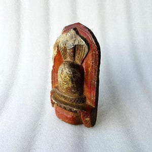 Puede incluir: Una peque&ntilde;a estatua de madera tallada de una deidad con un acabado de pintura roja, dorada y marr&oacute;n. La estatua tiene la forma de una persona con una capucha de cobra en la cabeza.