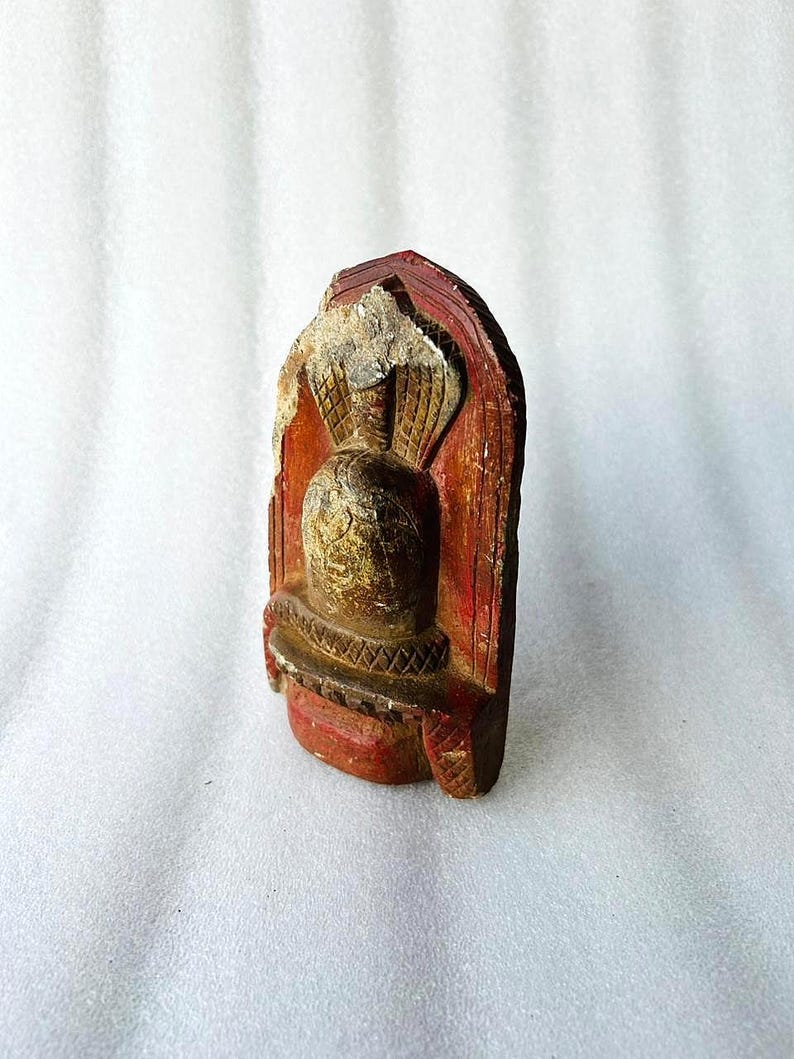 Puede incluir: Una peque&ntilde;a estatua de madera tallada de una deidad con un acabado de pintura roja, dorada y marr&oacute;n. La estatua tiene la forma de una persona con una capucha de cobra en la cabeza.