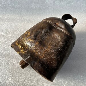 May include: A brown metal cowbell with an intricate engraved design. The bell has a wooden handle and a worn, aged appearance.