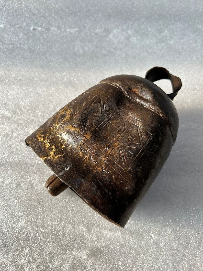 May include: A brown metal cowbell with an intricate engraved design. The bell has a wooden handle and a worn, aged appearance.