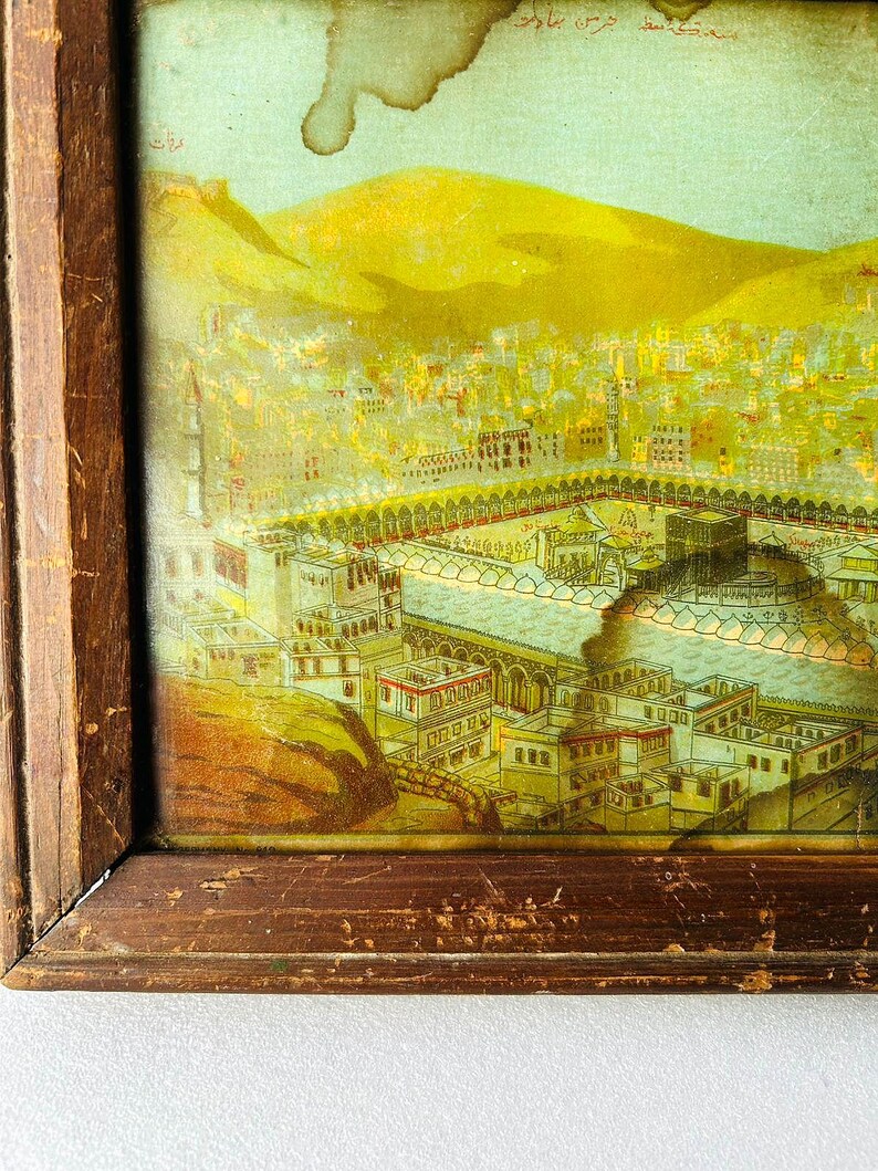 Puede incluir: Una ilustraci&oacute;n vintage enmarcada de la Kaaba en La Meca, Arabia Saudita. La imagen est&aacute; en un marco de madera y presenta una representaci&oacute;n detallada del sitio sagrado, rodeado de edificios y una colina en el fondo.