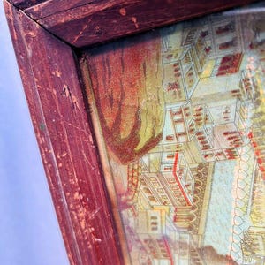 Puede incluir: Un marco de madera vintage con una impresi&oacute;n descolorida y colorida de una ciudad. El marco es rojo con un acabado desgastado y envejecido. La impresi&oacute;n representa edificios y un r&iacute;o con un puente.