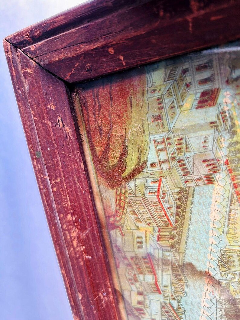 Puede incluir: Un marco de madera vintage con una impresi&oacute;n descolorida y colorida de una ciudad. El marco es rojo con un acabado desgastado y envejecido. La impresi&oacute;n representa edificios y un r&iacute;o con un puente.