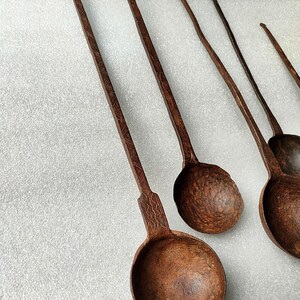 以下が含まれることがあります： 長い柄のついたアンティークの金属製のレードルが5つ。レードルは、さび付いた緑青のある濃い茶色の金属製。柄はシンプルな模様で飾られている。