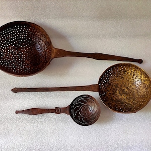 May include: Three antique metal strainers with long handles. The strainers are made of a dark brown metal and have a circular shape with holes throughout. The largest strainer has a diameter of approximately 10 inches.