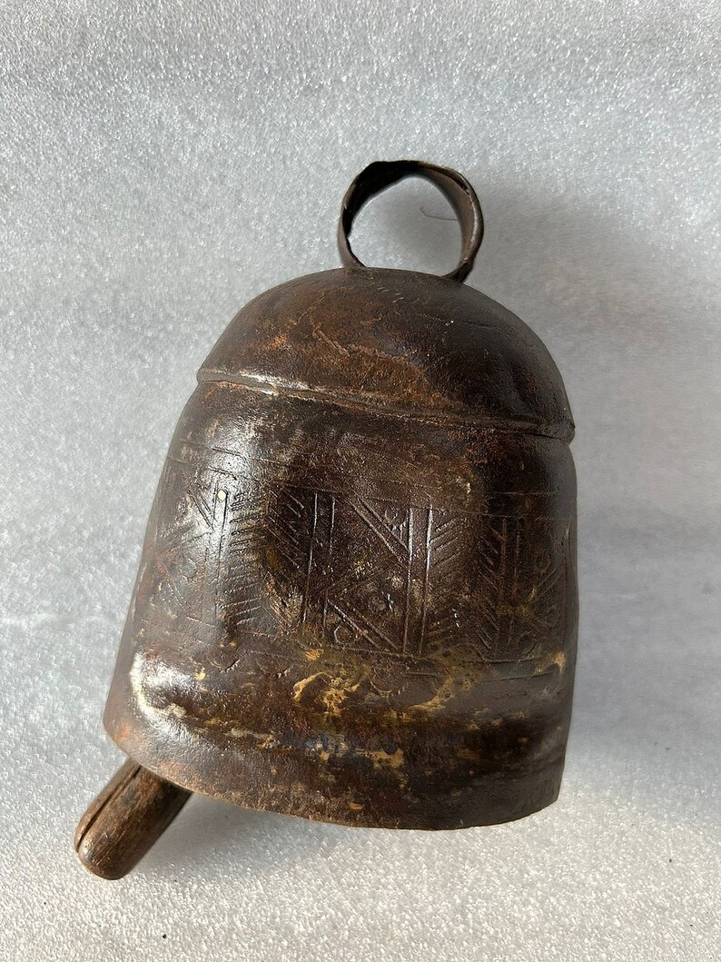 May include: A brown metal bell with a wooden handle and a decorative etched pattern. The bell has a ring at the top for hanging.