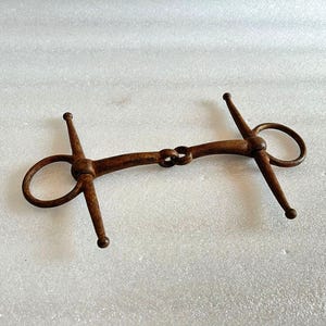 Pode incluir: Um freio de cavalo de metal enferrujado com dois anéis e uma junta central. O freio é projetado para ser usado na boca de um cavalo.