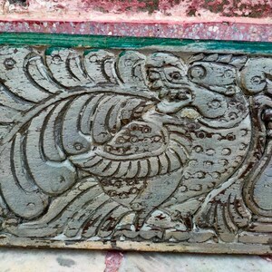May include: A close-up of a carved wooden panel with a detailed design of a mythical creature with wings and a beak. The panel is painted in a light gray color and has a weathered appearance.
