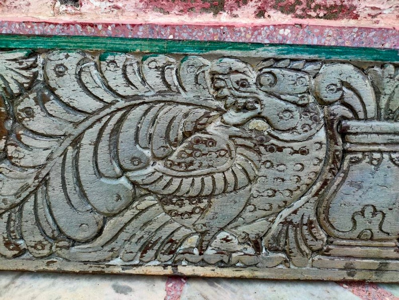 May include: A close-up of a carved wooden panel with a detailed design of a mythical creature with wings and a beak. The panel is painted in a light gray color and has a weathered appearance.