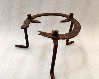 May include: A rusty, three-legged metal tripod stand with a circular top. The stand has a weathered appearance, suggesting age and use. The legs are bent and angled, providing stability. The top has three curved supports.