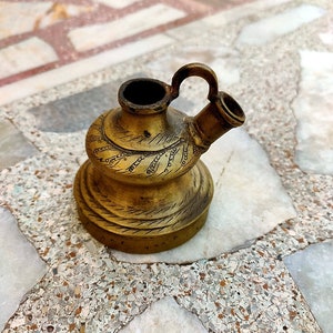 May include: A small, ornate brass teapot with a decorative handle and intricate engravings. The teapot is sitting on a tiled surface.
