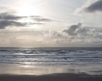 May include: A seascape with a cloudy sky and a sandy beach with waves crashing on the shore.