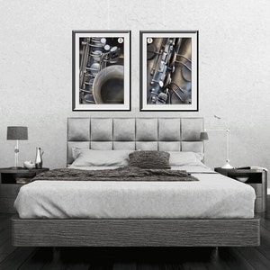 May include: Two framed black and white photographs of a saxophone. The photographs are hanging on a white wall above a gray upholstered bed with a gray headboard. The bed is in a bedroom with a gray floor and two decorative vases on the floor.