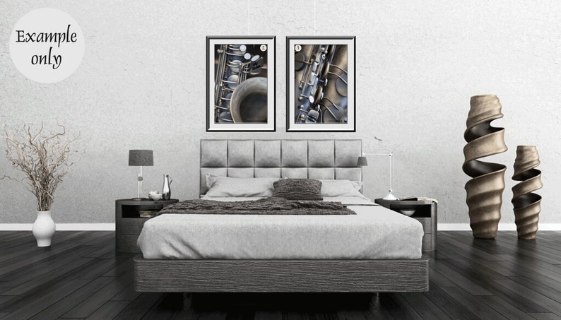 May include: Two framed black and white photographs of a saxophone. The photographs are hanging on a white wall above a gray upholstered bed with a gray headboard. The bed is in a bedroom with a gray floor and two decorative vases on the floor.