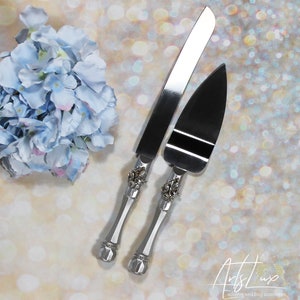 May include: A set of two silver cake knives with ornate handles and rhinestones. The knives are on a sparkly background.